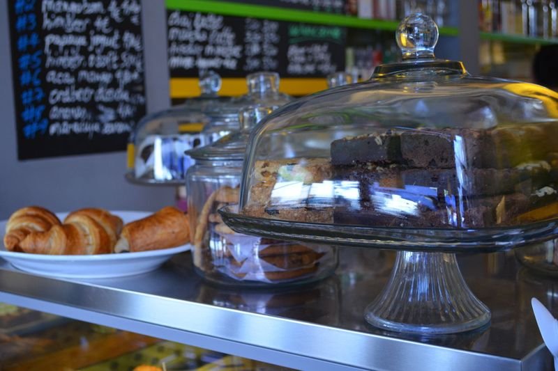 Handgeschriebene Tafeln mit Kaffeespezialitäten über der Kuchentheke, davor Croissants unter Glasglocken
