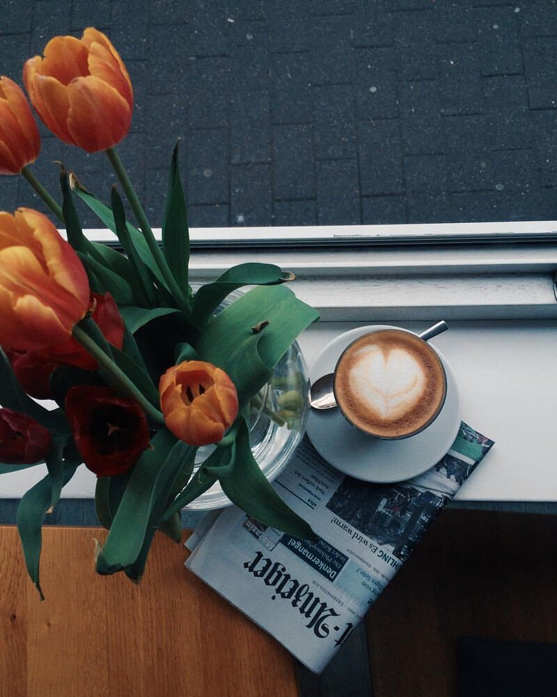Cappuccino und Tulpen auf einem Tisch am Fenster, daneben eine Zeitung